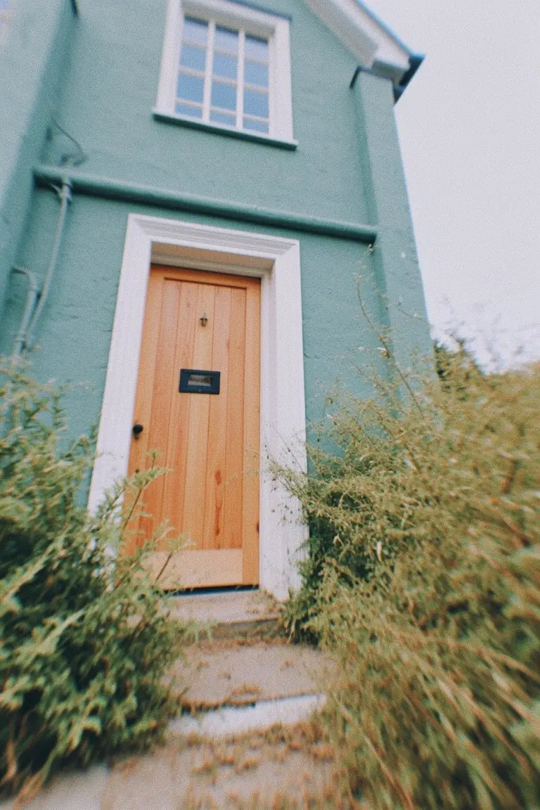 Teal house exterior with white trim framing a wooden door amid overgrown greenery