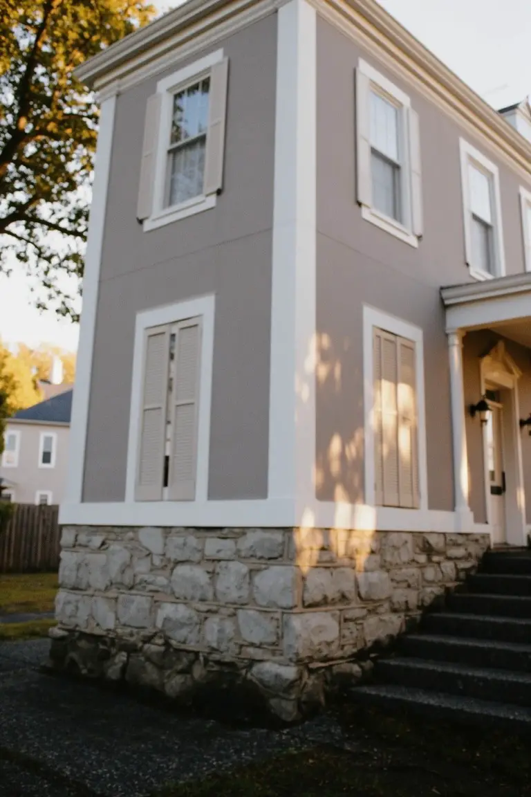 Corner view of a classic house painted soft greige with white trim, stone base, and shutters