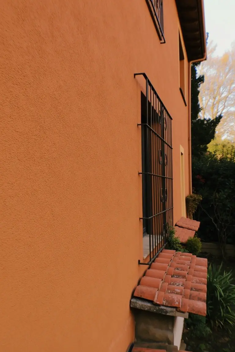 Warm terracotta stucco house wall with black wrought iron window bars and clay tile roof ledge