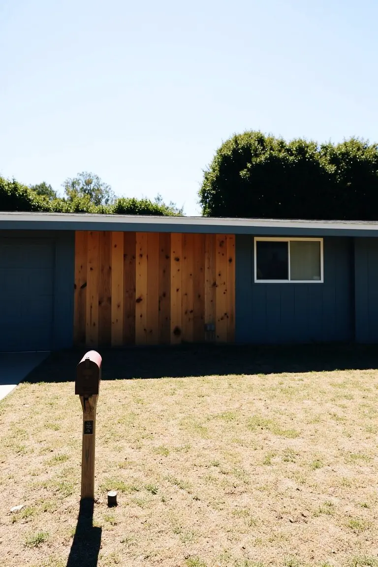 Mid-century modern house with deep navy blue walls accented by vertical cedar planks, white window trim, and a red mailbox on a dry lawn