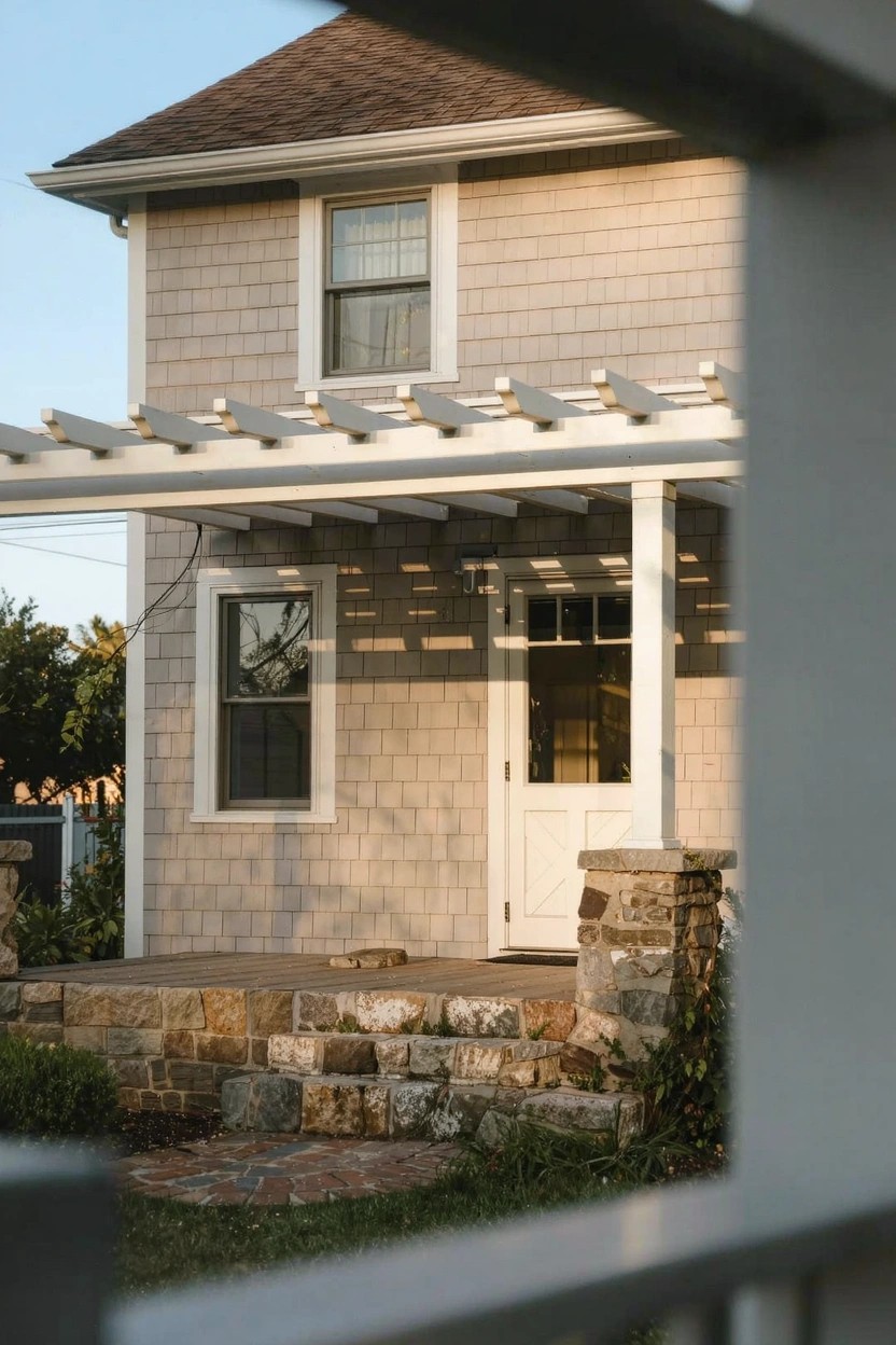 Light greige shingled house exterior with white pergola, door, and stone steps under a blue sky