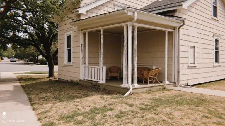 Two-story house with warm beige siding, covered front porch, wicker chairs, and wood accents under leafy trees