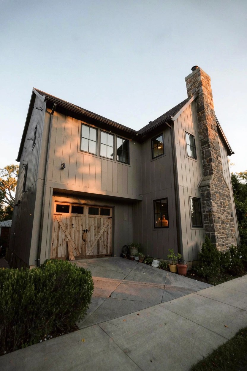 Modern farmhouse exterior with warm greige siding, wood garage doors, and stone chimney at dusk