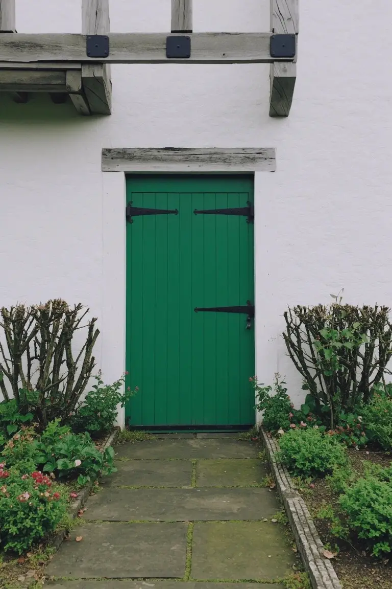 A deep green wooden front door centered on white cottage walls, with black iron hardware, timber lintel above, and garden shrubs on either side of a stone path