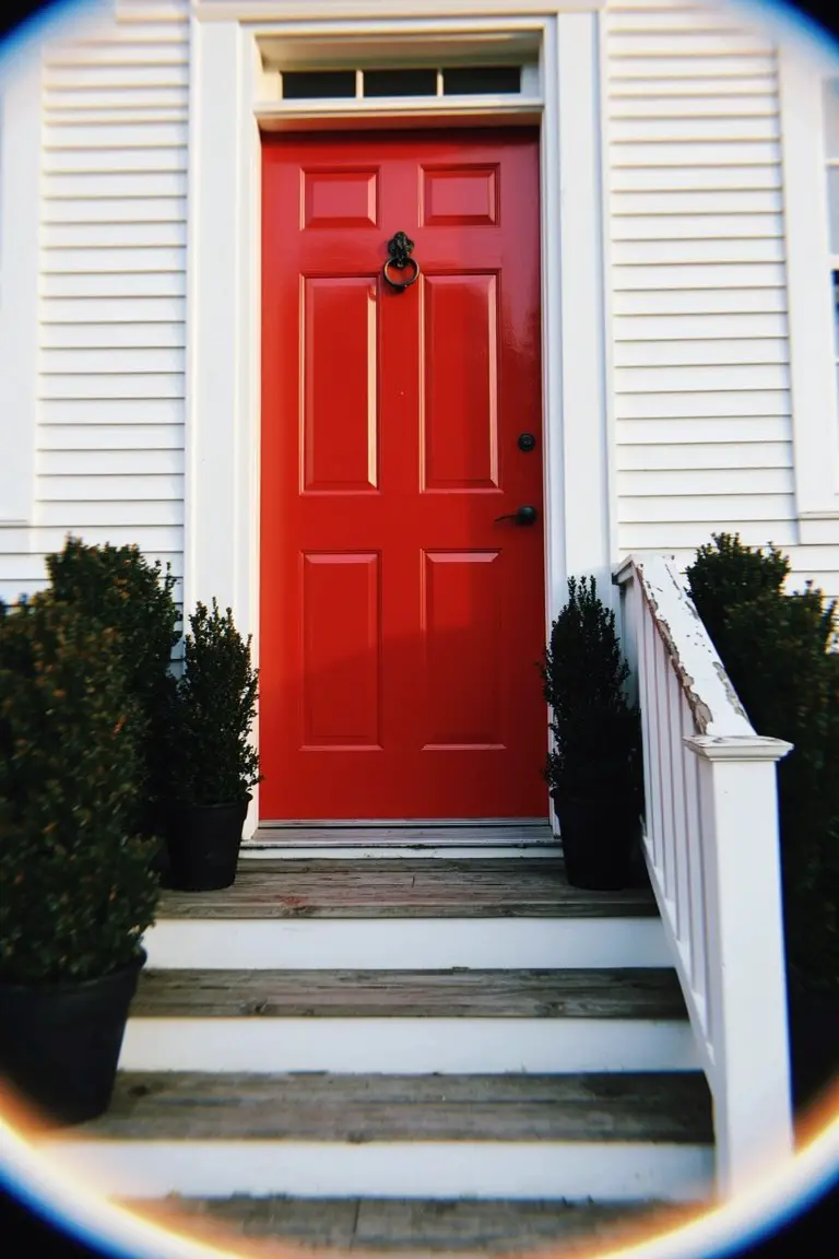 Glossy bright red front door centered on white clapboard house with boxwood shrubs and wood steps