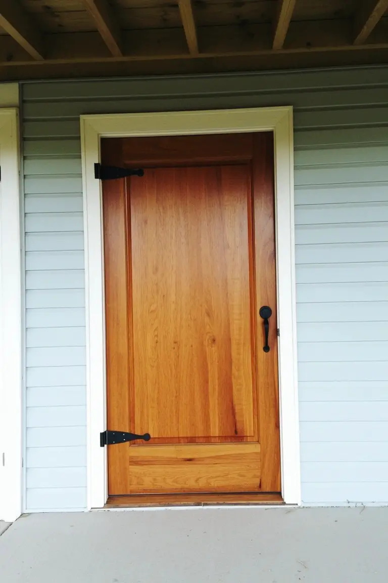 Natural golden brown wood front door with black iron hardware and white trim on light blue clapboard siding