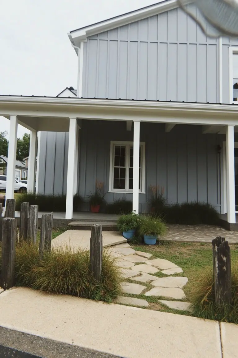 Light cool gray board-and-batten siding on a farmhouse-style house with white trim, wood porch posts, and a stone walkway edged in grasses