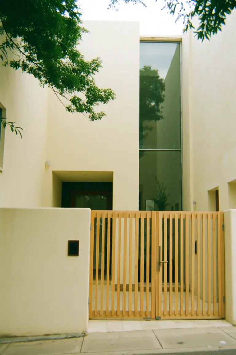 Small modern home exterior in pale warm beige with wooden slat gate and tall glass window under tree shade