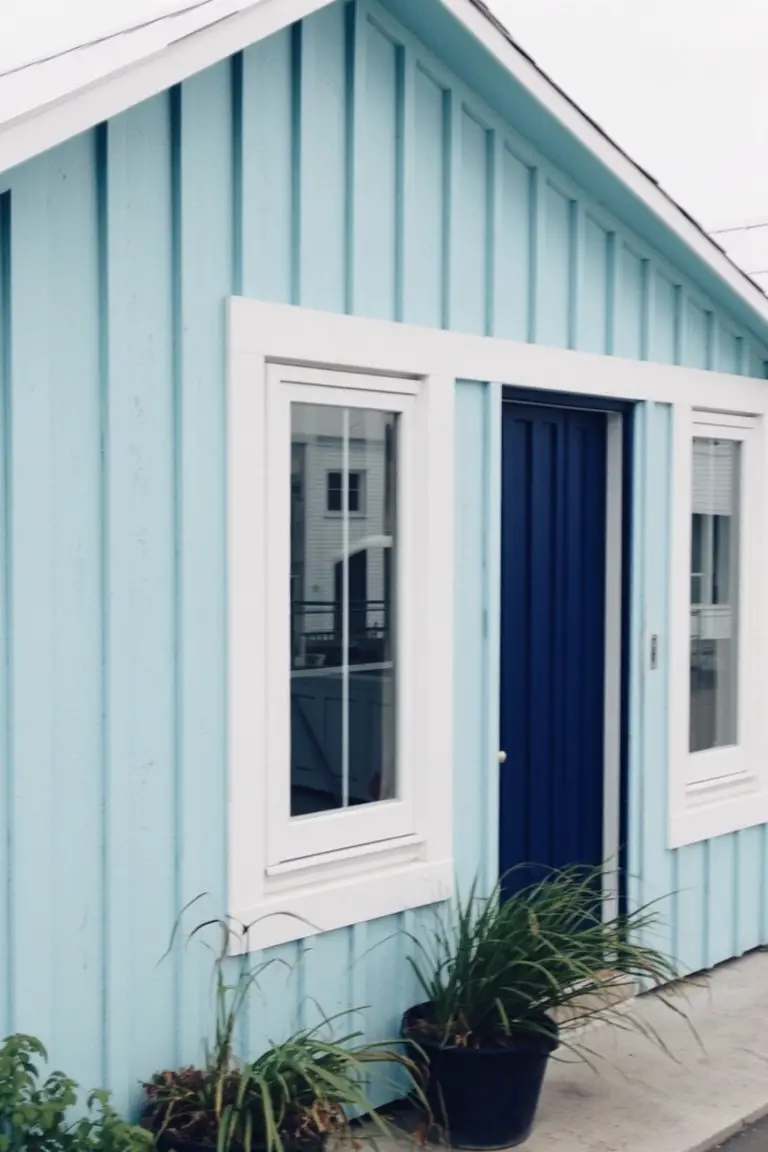 Small house with light aqua blue vertical siding, white window trim, navy blue front door, and potted plants by the steps