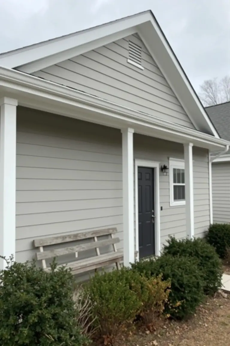 Small home exterior with soft light gray siding, white porch columns, dark blue door, and wooden bench