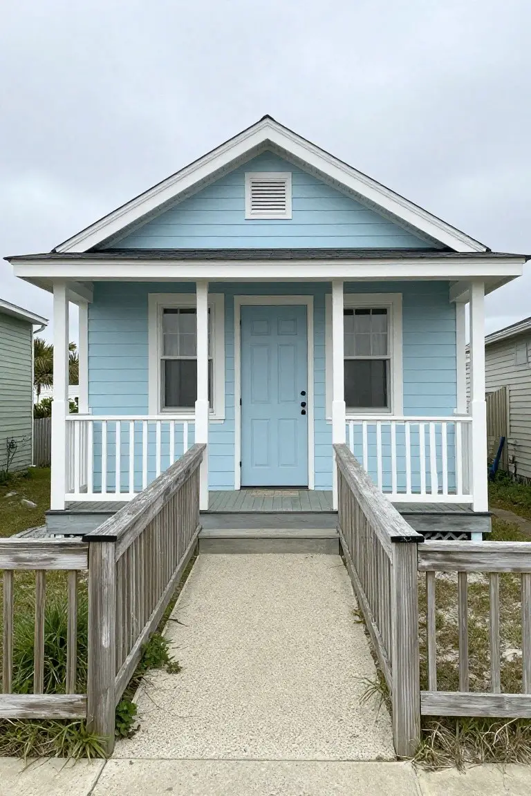 Light blue cottage-style house exterior with white trim, front porch, and wood railing on an elevated lot