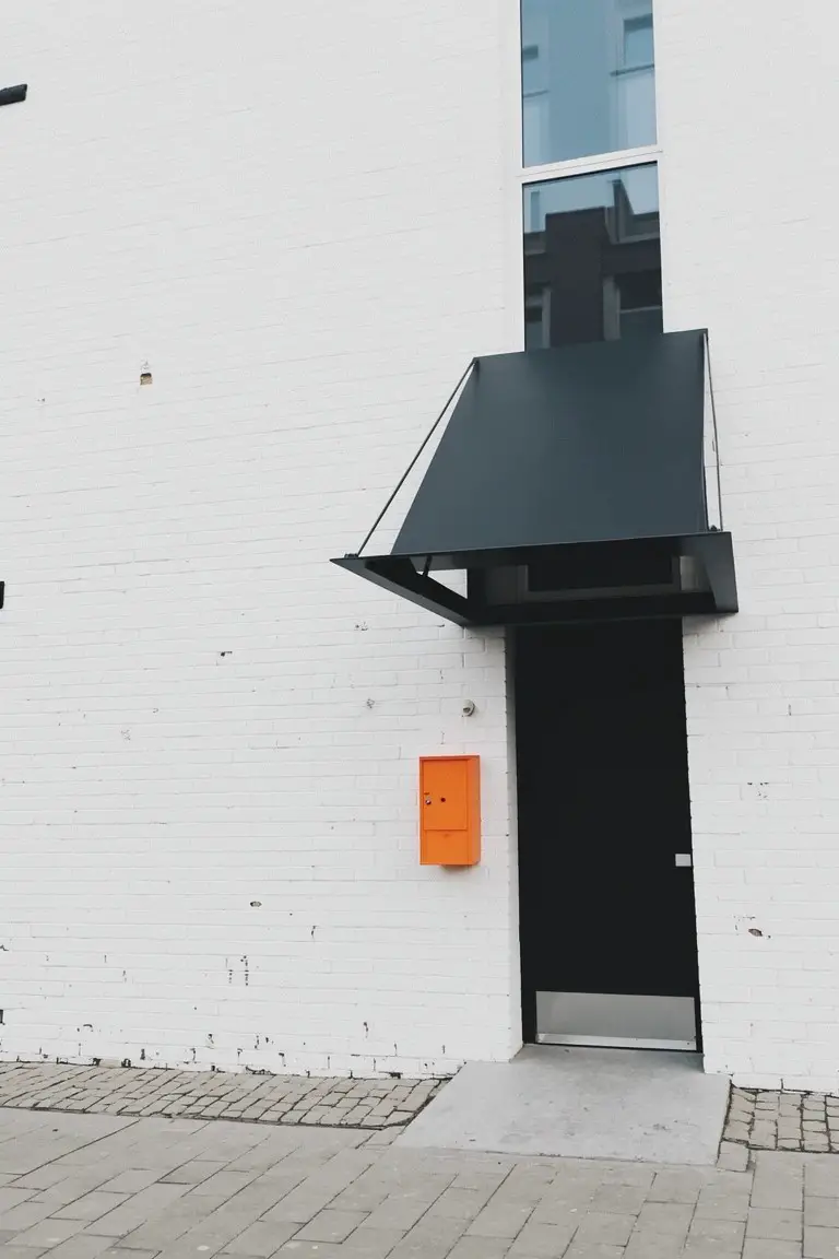 Crisp white exterior wall with black door, dark green awning, and orange mailbox on brick house