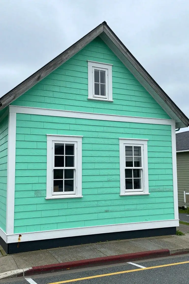 Small gabled house with turquoise clapboard siding, white trim, and black base on a sidewalk