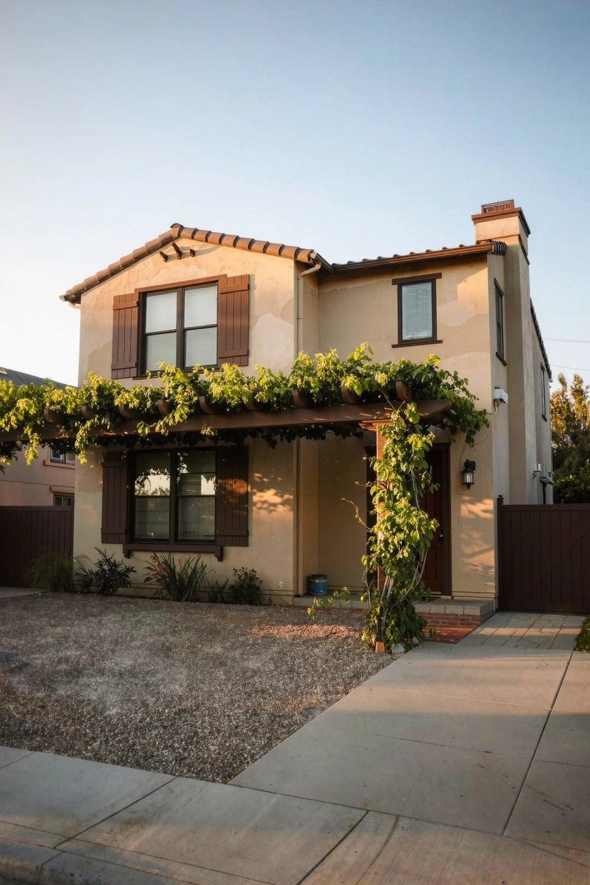 Warm beige stucco house exterior with brown wood shutters, grapevine-covered pergola, and gravel driveway