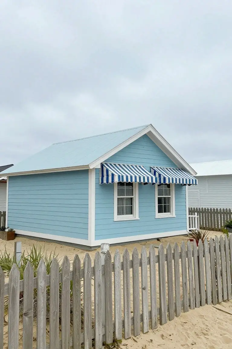 Light blue beach cottage with white trim, striped awnings, and picket fence