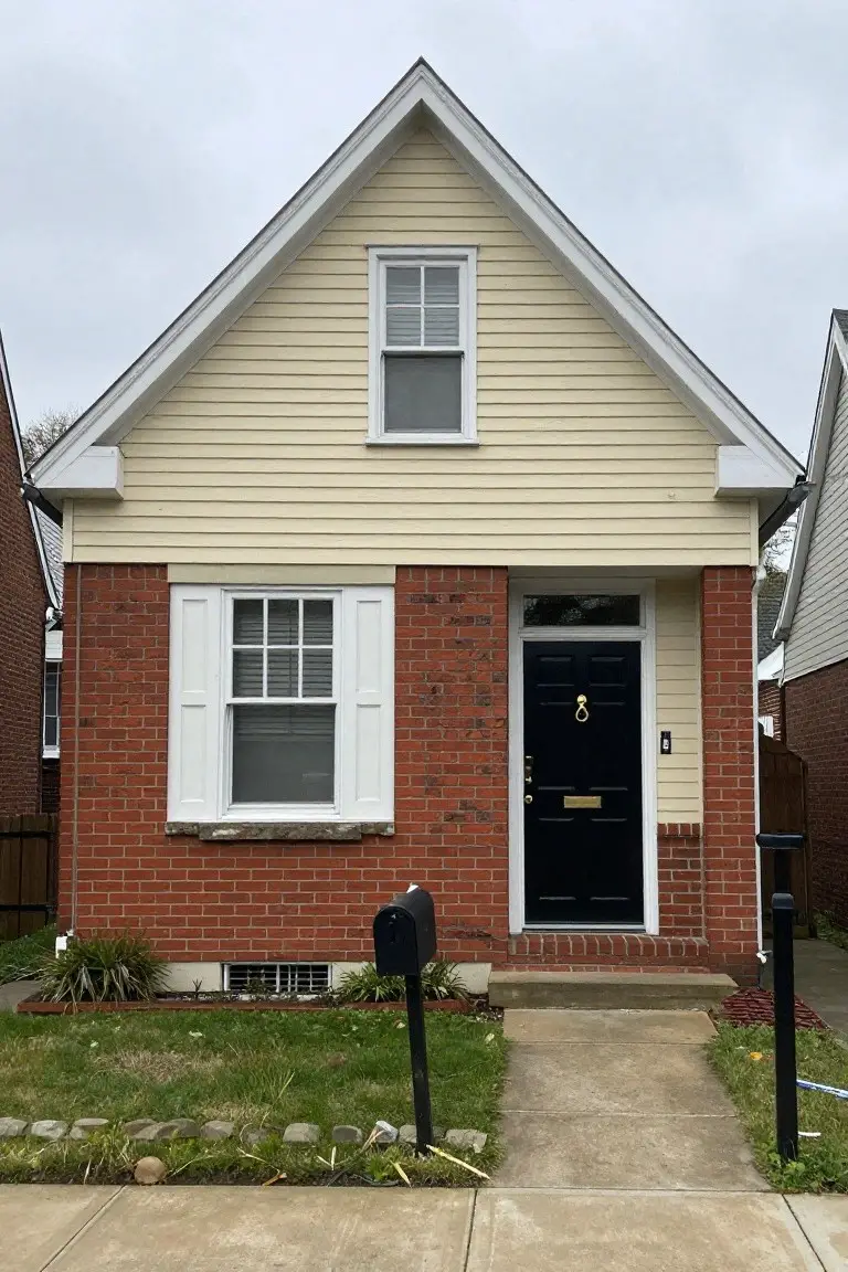 Bungalow exterior featuring warm beige siding over red brick foundation, white window trim, and black front door