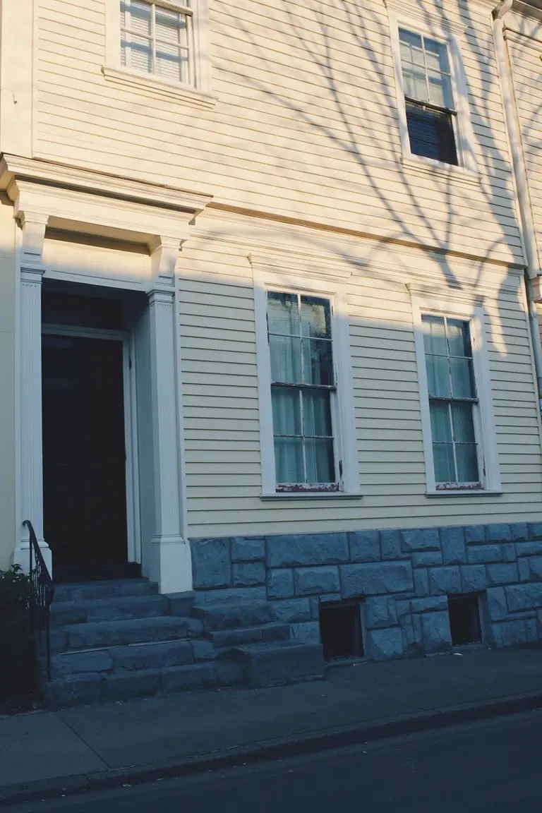 Two-story clapboard house with pale yellow siding, granite stone foundation, black front door, and white trim in late afternoon light