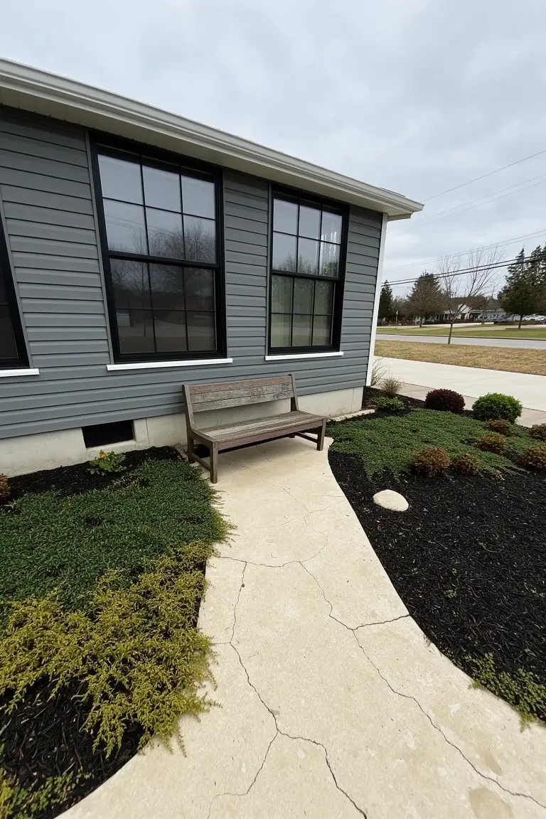 Ranch house exterior featuring cool gray siding, black-framed windows, wooden bench on a stone path, and low shrubs