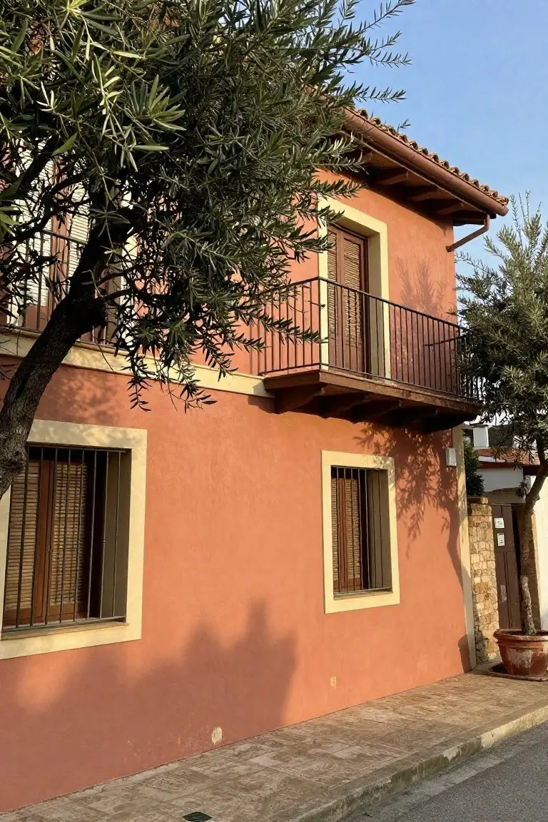 Two-story terracotta house with olive trees, wooden shutters, wrought-iron balcony, and stone accents under a tiled roof