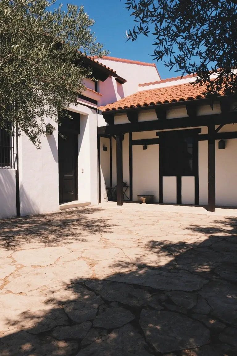 Crisp white stucco walls on a home exterior with terracotta tile roof, black wood beams, and olive trees casting shadows on stone pavers