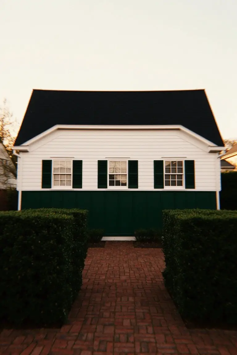 White house with deep green shutters and lower panel, black roof, brick pathway and hedges