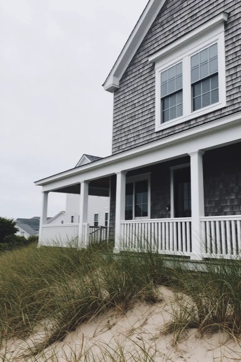 Coastal shingle-style home painted soft gray with bright white trim on porch columns and railing, beach grass nearby