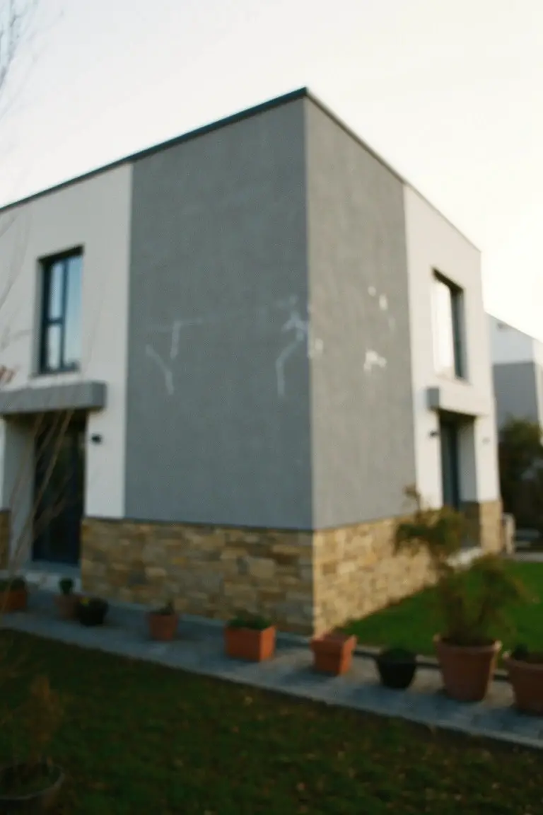 Modern house corner with polished cool gray walls, white trim accents, beige stone base, and potted plants in front yard