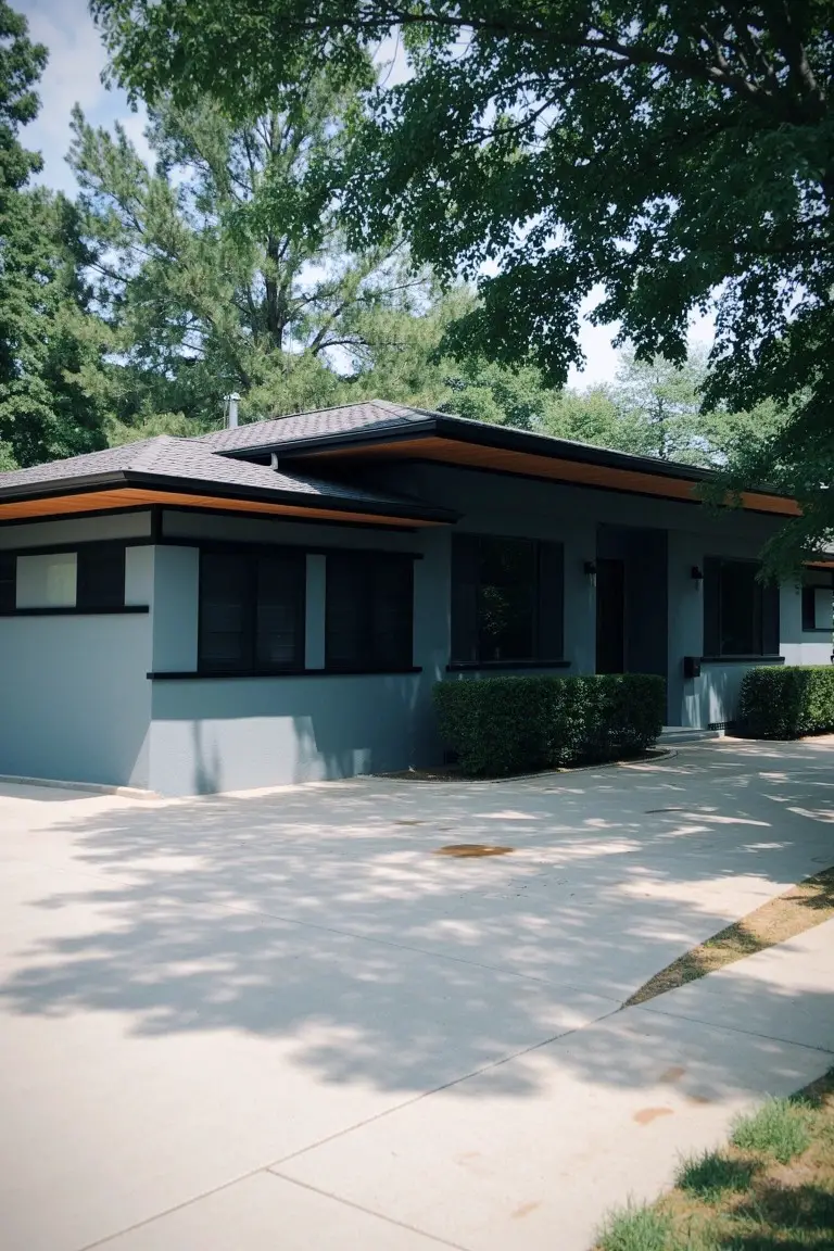 Modern house exterior in light cool gray with black windows, orange trim accents, and a paved driveway under trees