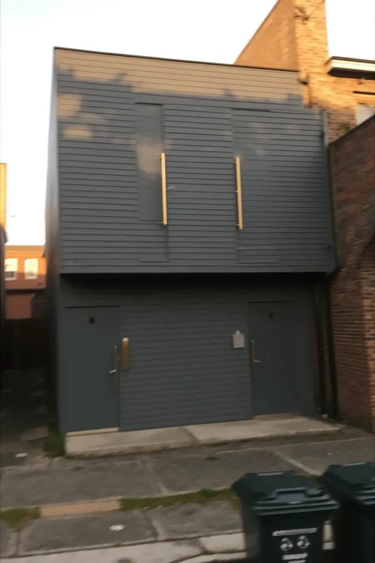 Narrow modern house with polished dark gray vertical siding, brick side wall, and slim gold door handles against a paved alley