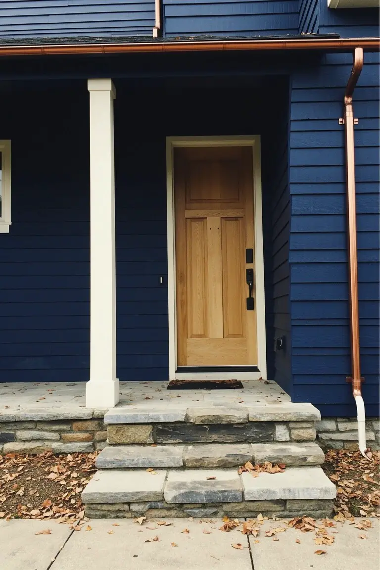Deep navy blue house siding paired with white porch columns, oak door, and stone steps