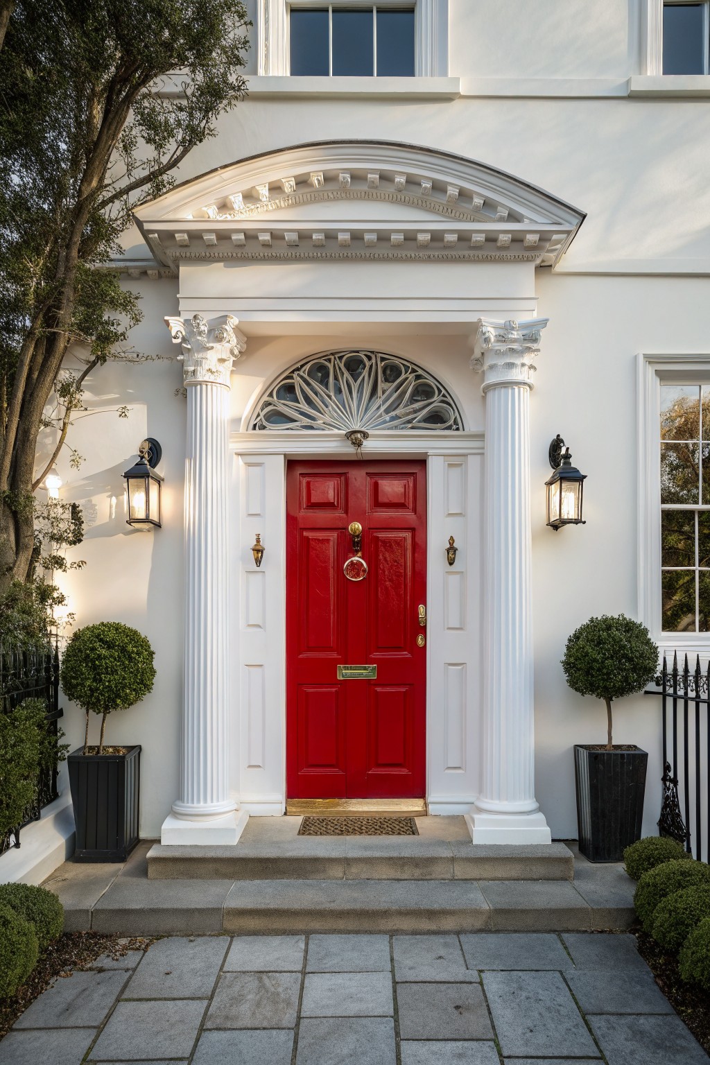 White stucco house facade with red front door centered under Ionic columns and arched fanlight window, flanked by lanterns and black pots of clipped boxwoods on stone steps.