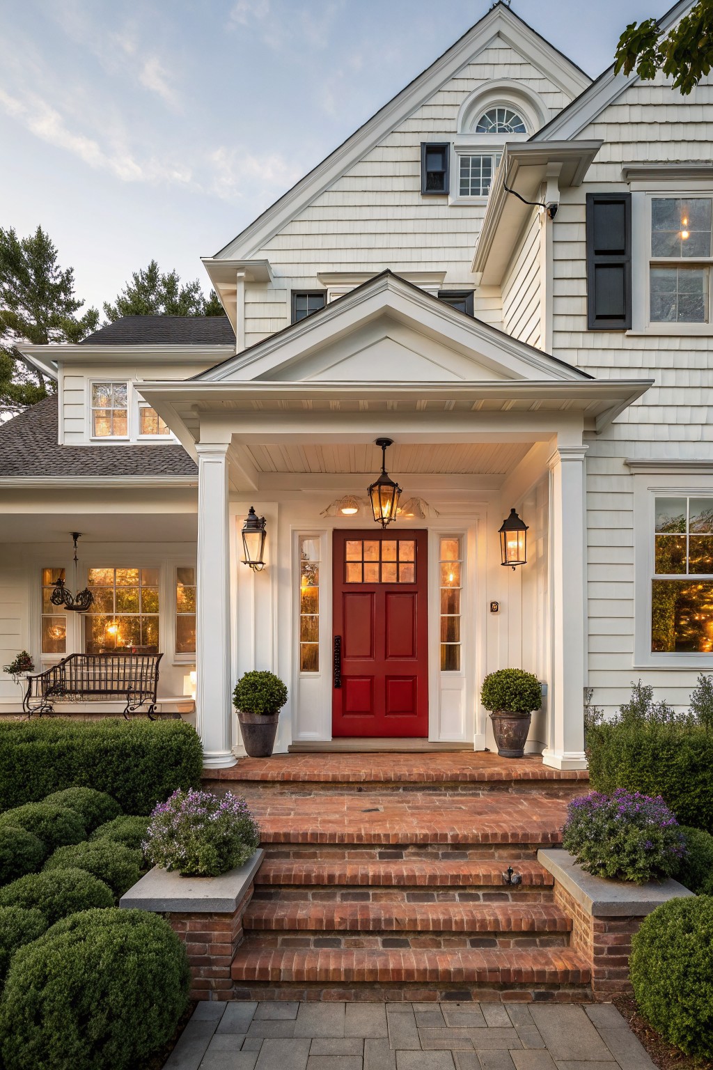 White shingle-style house exterior featuring a bright red double front door under a columned porch, flanked by lanterns, with brick steps leading up from a stone-edged path and boxwood shrubs.