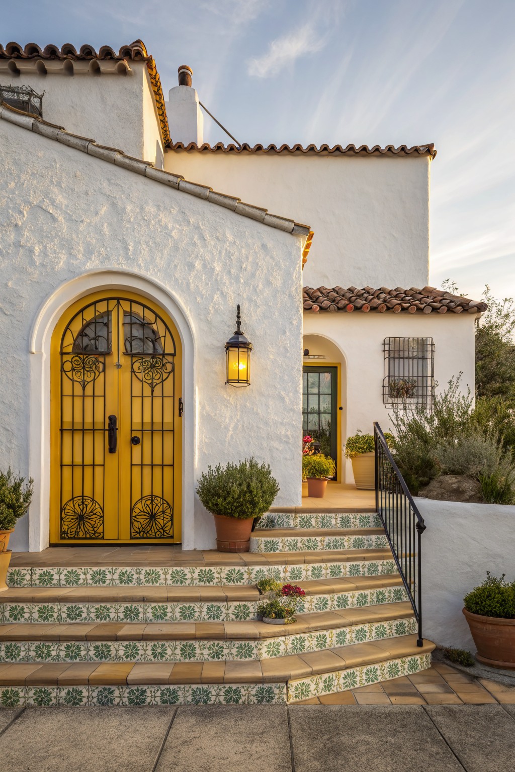 White stucco house exterior featuring a tall arched yellow wrought-iron front door with black grillework, flanked by potted plants and a lantern light, with green-and-white tiled steps leading up to it.