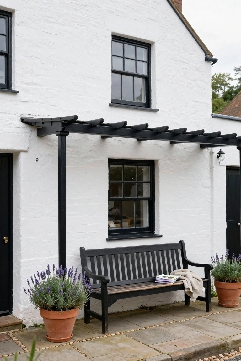 White rendered cottage exterior with black window frames, front door, metal pergola supported by posts, wooden bench with throw blanket and books, lavender in terracotta pots on a paved patio with pebble edging.