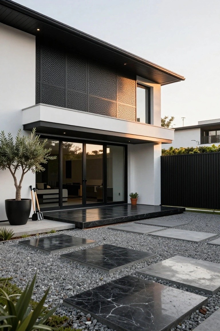 Modern white stucco house exterior featuring black perforated metal screens on upper walls, large black-framed sliding glass doors, entry platform with black marble steps, gravel yard with stone pavers, potted olive tree, and agave plants.