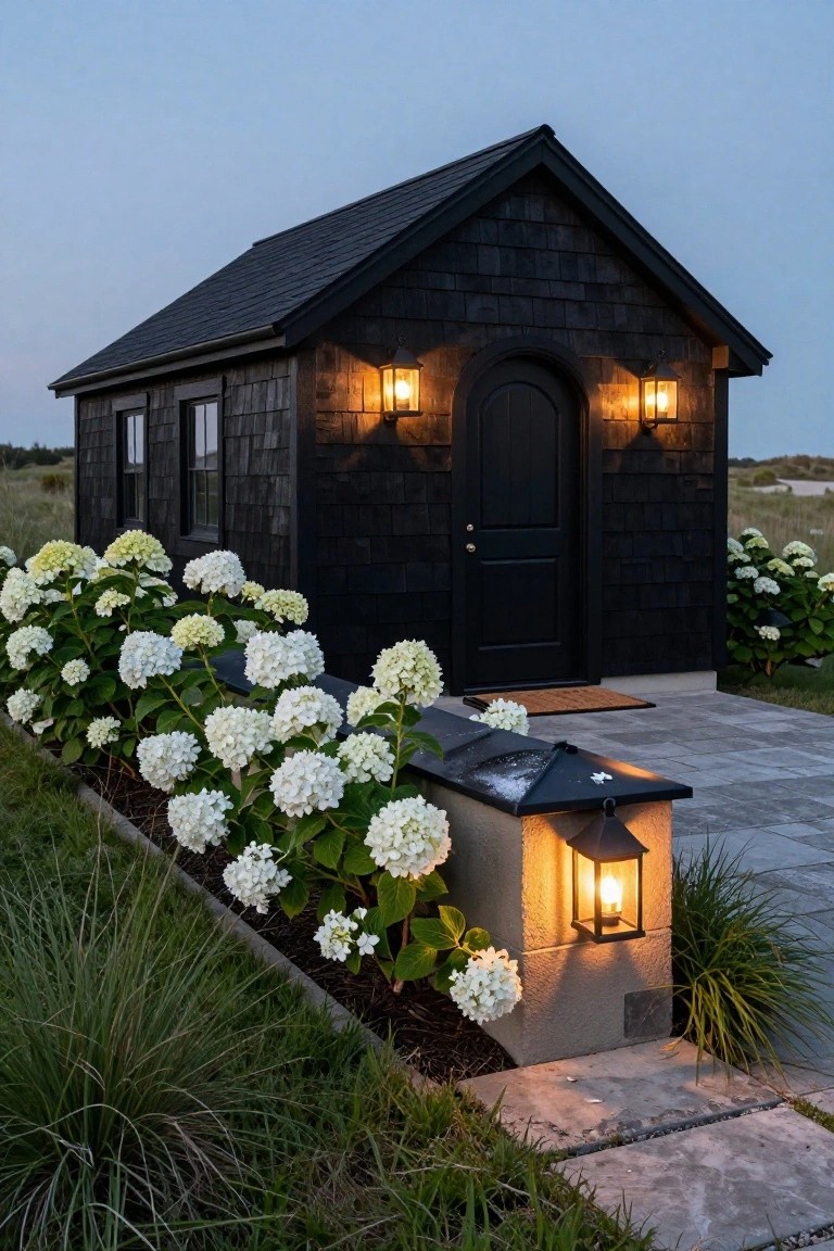 Small black shingled house exterior at dusk with warm lantern lights flanking the arched black door and a post lantern nearby, bordered by white hydrangea bushes and dune grass.