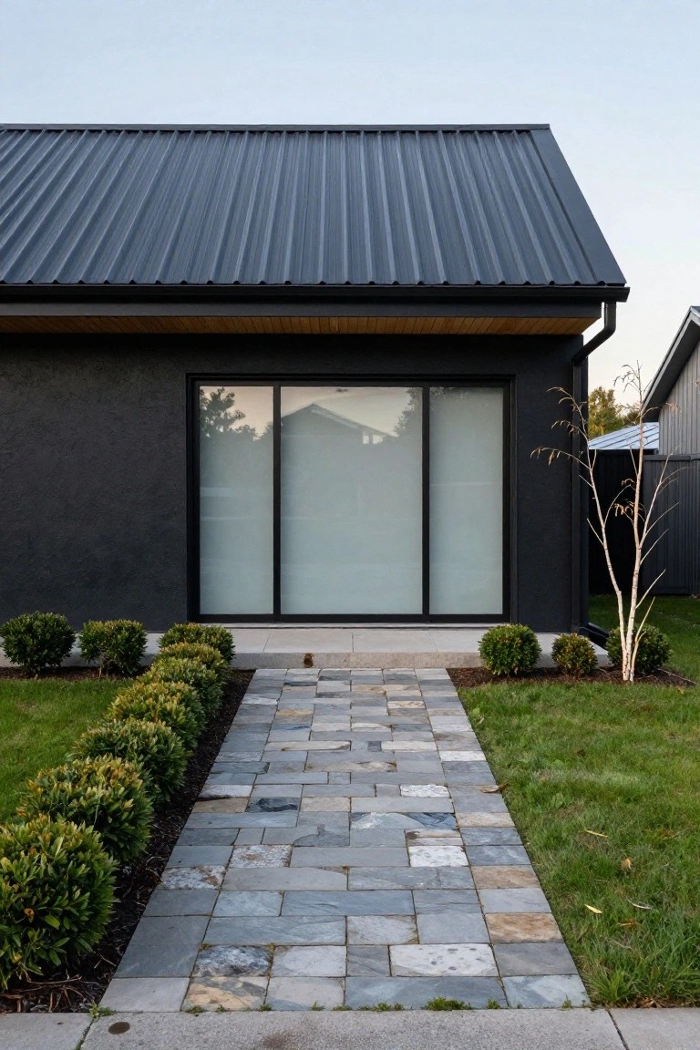 Front view of a modern single-story black house with dark corrugated metal gable roof, black-framed frosted glass sliding doors, wood soffit overhang, stone paver pathway lined with boxwood shrubs, and a white birch tree beside the entry.