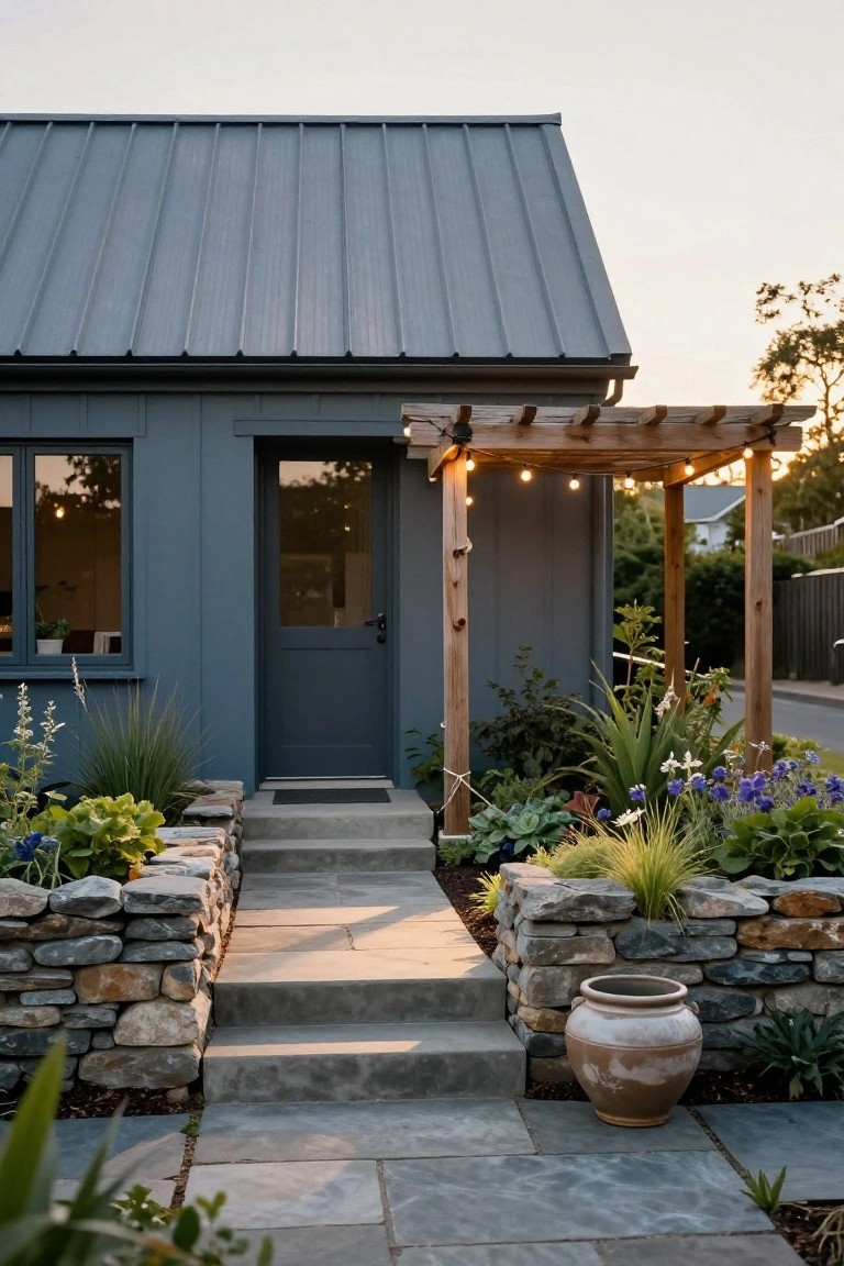 Small dark blue house with gray metal roof and wooden pergola strung with lights over dark blue glass door, concrete steps flanked by stone walls and drought-tolerant plants.