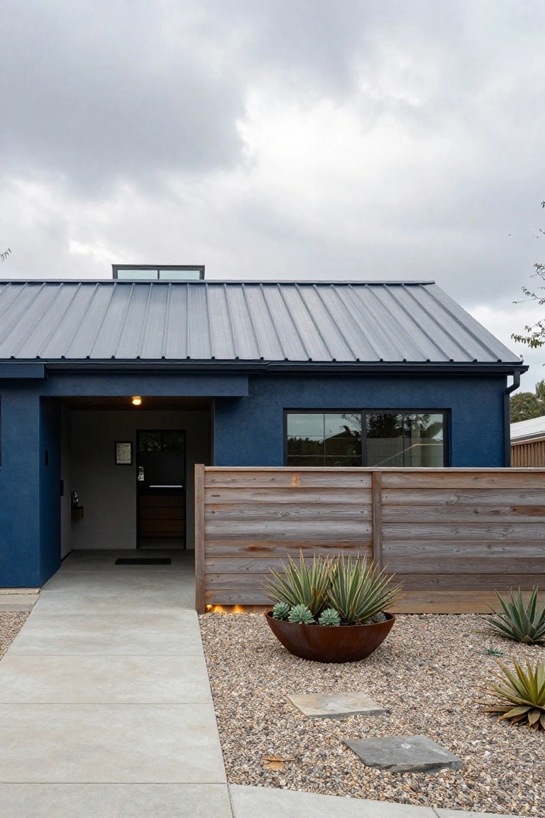 Front view of a deep blue stucco house with dark standing seam metal roof, recessed open entryway with wood slat fence, concrete walkway, gravel yard planted with agave succulents, and a large terracotta pot.