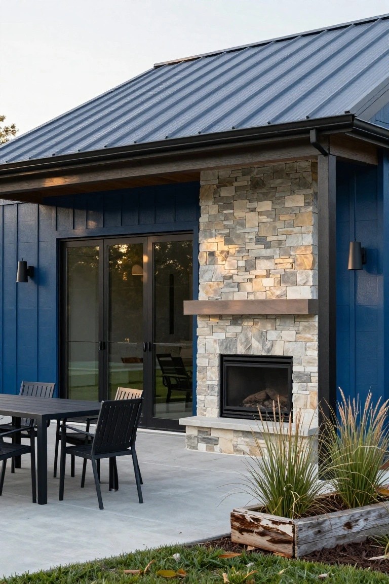 Blue board-and-batten siding on house exterior with blue standing seam metal roof, tall stacked stone outdoor fireplace next to black sliding glass doors, concrete patio with black dining table and chairs, potted grasses nearby