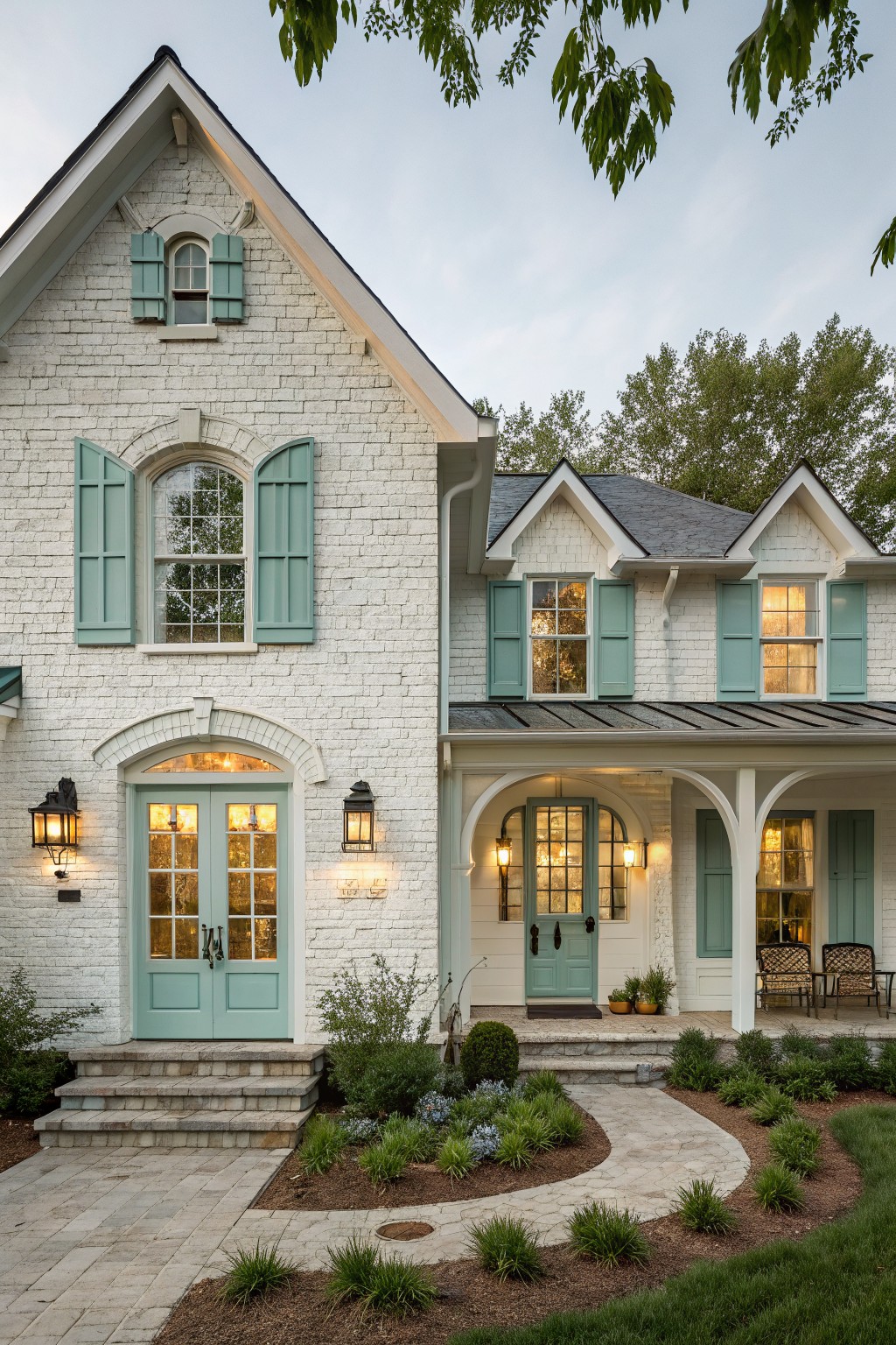 White brick two-story house with teal blue shutters on windows and doors, arched covered porch with lanterns, stone steps and curved paver pathway, and low plantings in the front yard.