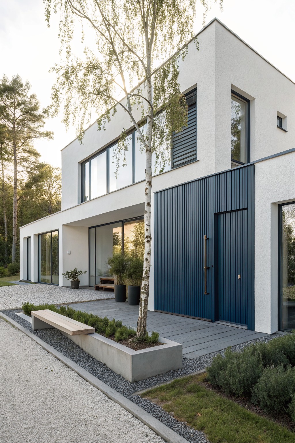 Modern white rectangular house exterior with navy blue vertical cladding on the entry door and adjacent wall, large glass windows, birch tree by entrance, gravel driveway, concrete planters, and surrounding landscaping.