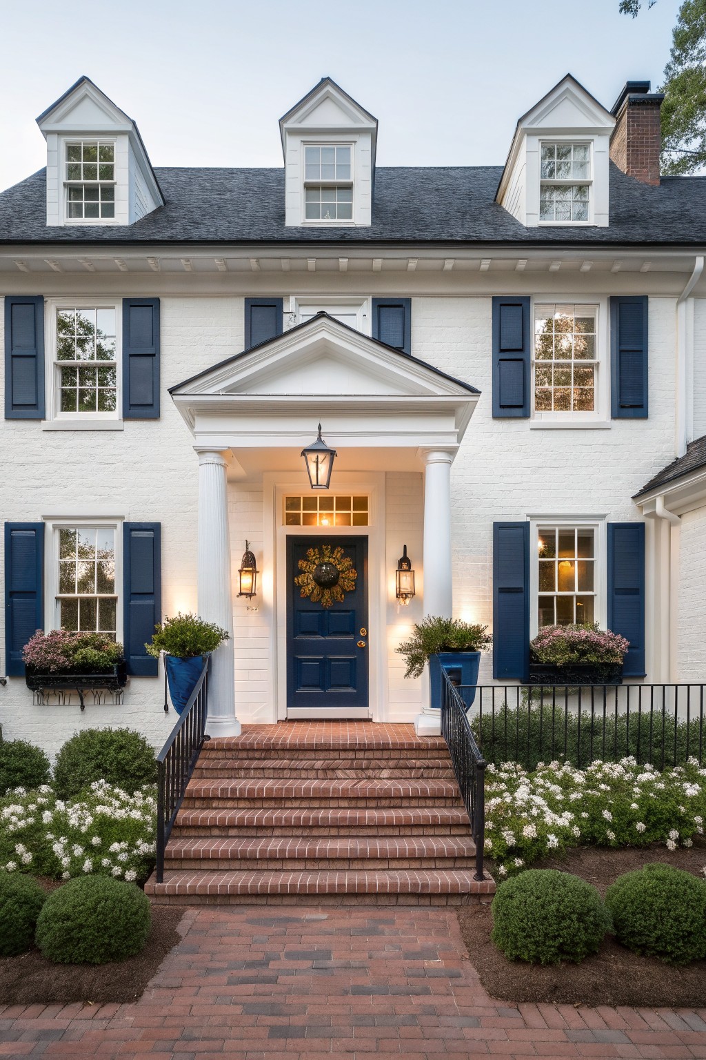 Two-story white clapboard house with dark gray roof, dormer windows, navy blue shutters on multiple windows, navy blue front door with brass knocker and fall wreath, white portico supported by fluted columns, wall lanterns, brick steps with black metal railing, boxwood shrubs, white flowers, and blue planters at entry.