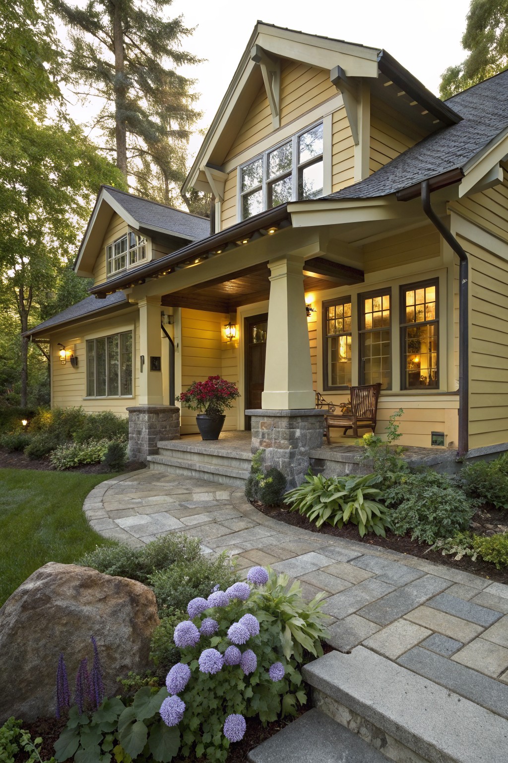 A pale yellow Craftsman-style house with dark shingled roof sections, covered front porch supported by stone pillars, potted red flowers on the porch, and a curving paver path through landscaped beds with purple hydrangeas and a large boulder.