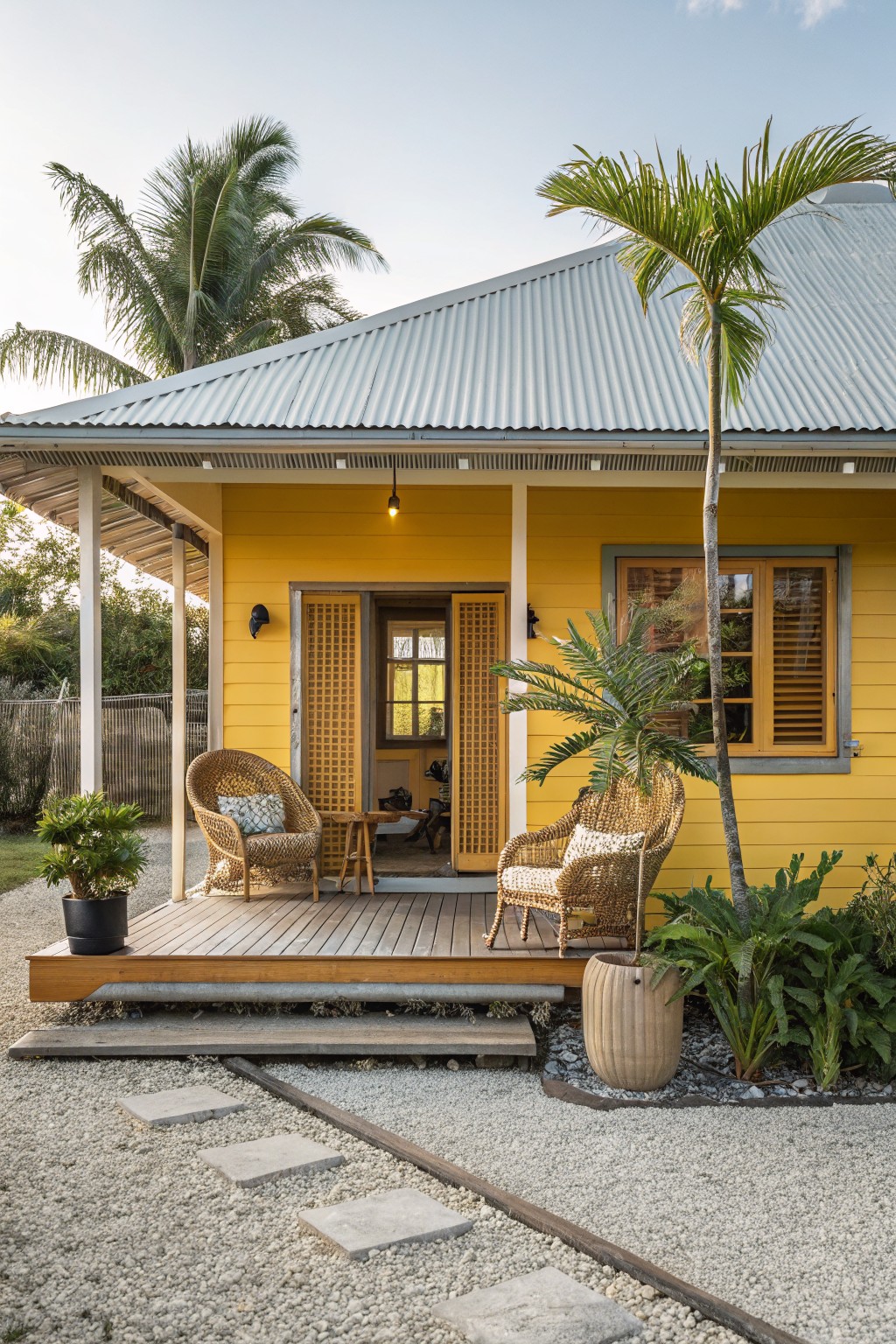A pale yellow weatherboard house with corrugated metal roof and covered porch holding two rattan chairs, flanked by palm trees and tropical plants on a gravel yard with stone path.