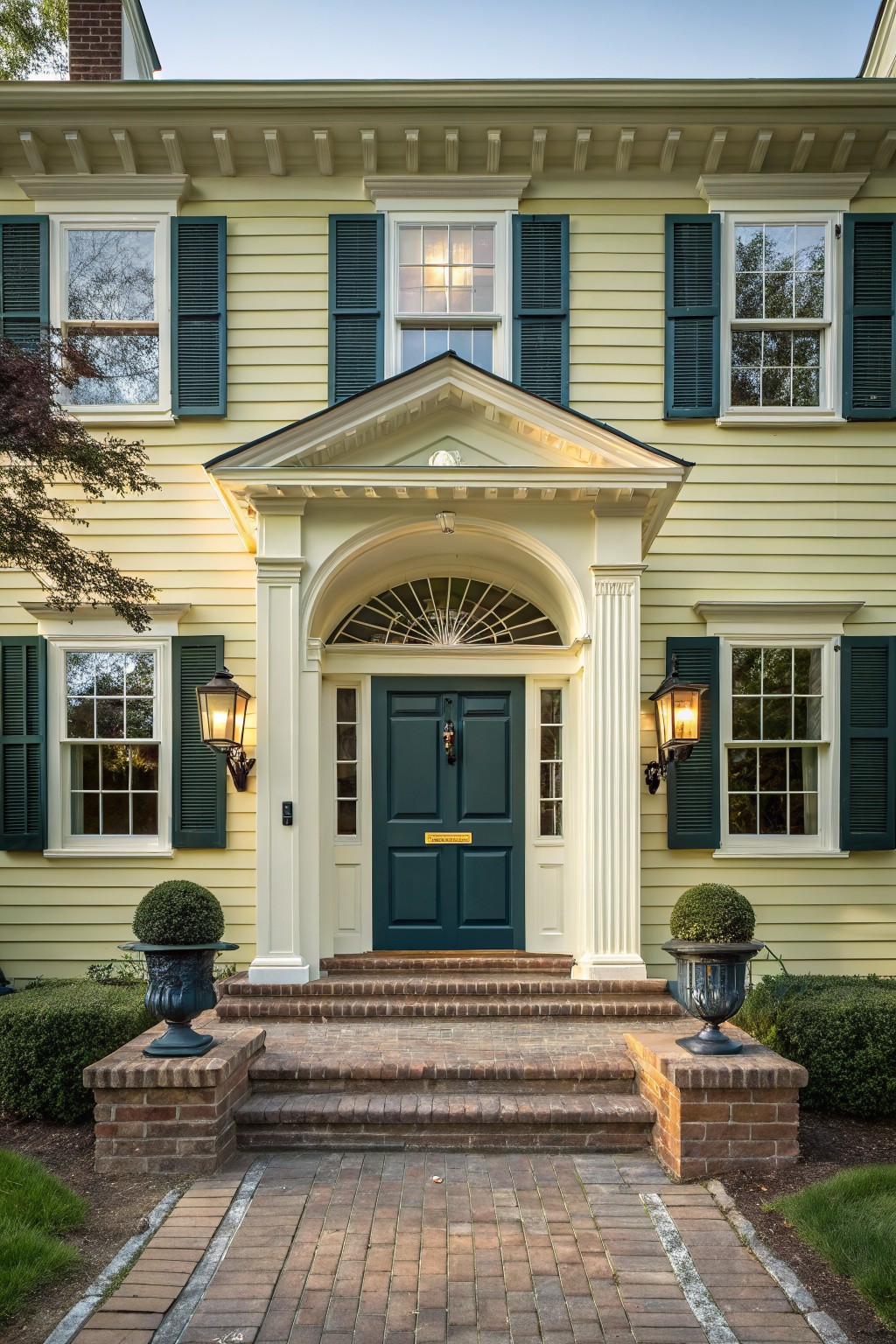 Pale yellow clapboard house with teal shutters, white pedimented portico supported by columns, dark green front door, lanterns, brick steps, urns with boxwood topiaries, and brick pathway.
