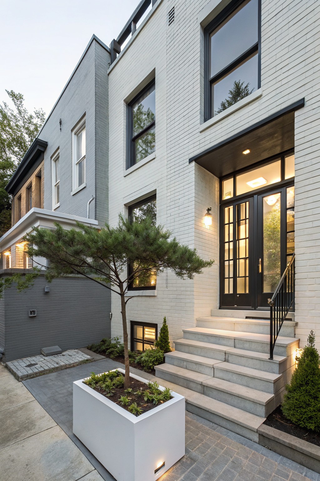 White brick rowhouse exterior with black-framed windows and double front doors, gray neighboring buildings, stone entry steps with black railing, a potted pine tree, and low plantings along the walkway.