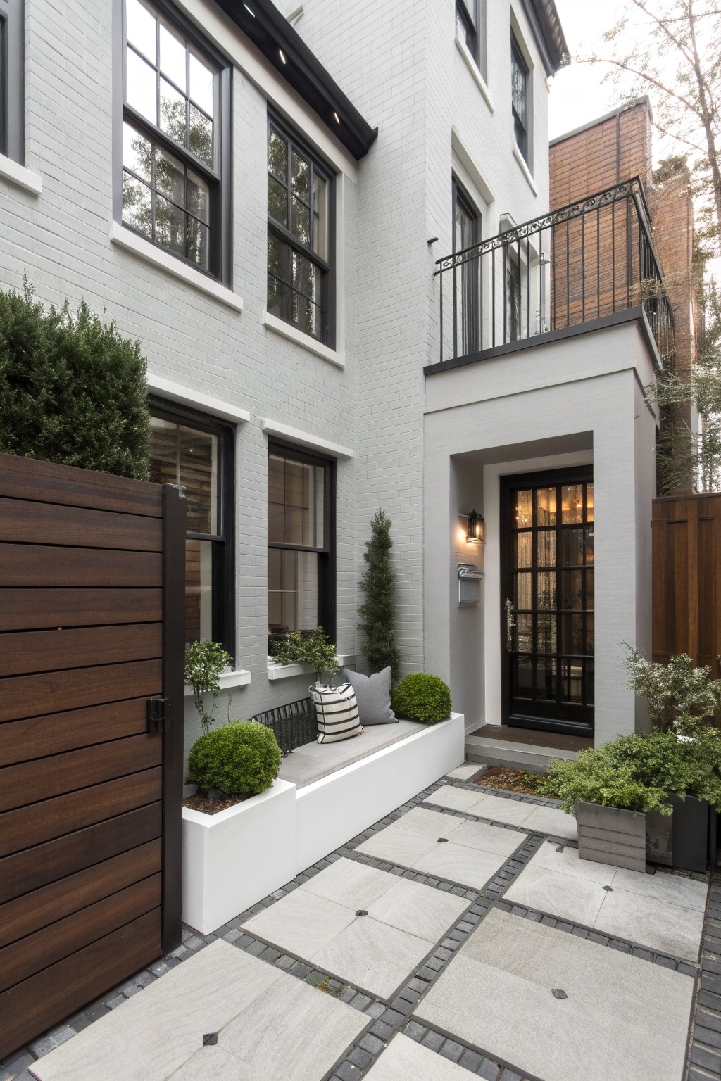 Light gray brick house exterior with black-framed windows and glass entry door, wooden privacy gate, paved stone pathway, built-in white bench with cushions and pillows, and potted shrubs along the entry wall.