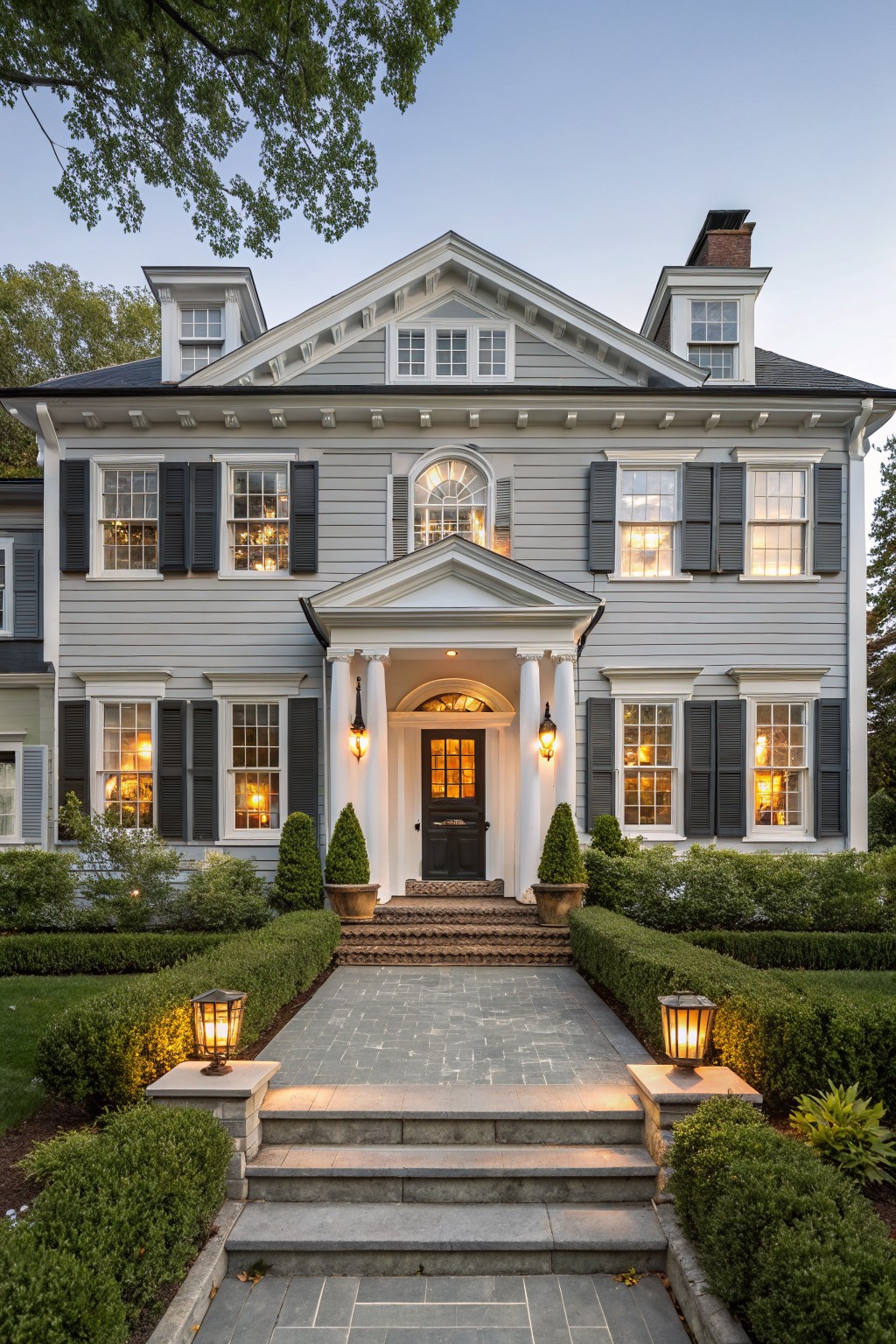 Two-story gray clapboard house with white trim, black shutters, columned white portico entry, black door, stone steps and pathway with lanterns, boxwood hedges, at dusk with interior lights on.