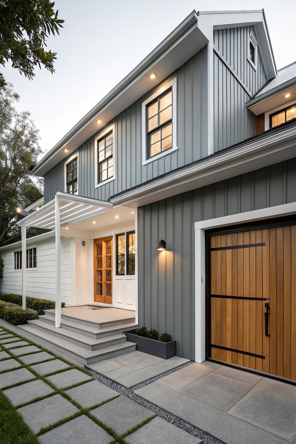 Two-story house exterior with gray vertical board-and-batten siding, white trim on windows and roofline, wooden garage door, white-covered entry porch with steps, and concrete paver pathway edged in grass.
